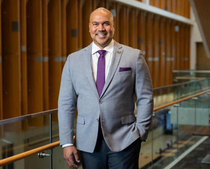 Dr. Bernard Rousseau in a light-gray suit with a purple tie and matching pocket square stands beside a glass railing in a modern atrium lined with tall wooden panels