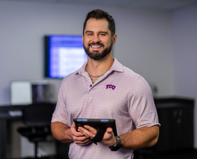 A smiling man wearing a light purple TCU polo shirt stands in a clinical research or laboratory setting, holding a tablet with both hands. He has short dark hair, a beard, and wears a watch and necklace. The background is softly blurred, showing computer workstations and monitors, emphasizing the professional healthcare and research environment.
