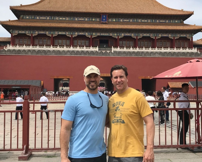 Two men pose together in front of the Hall of Supreme Harmony at the Forbidden City in Beijing, China, with visitors and traditional Chinese architecture visible in the background.